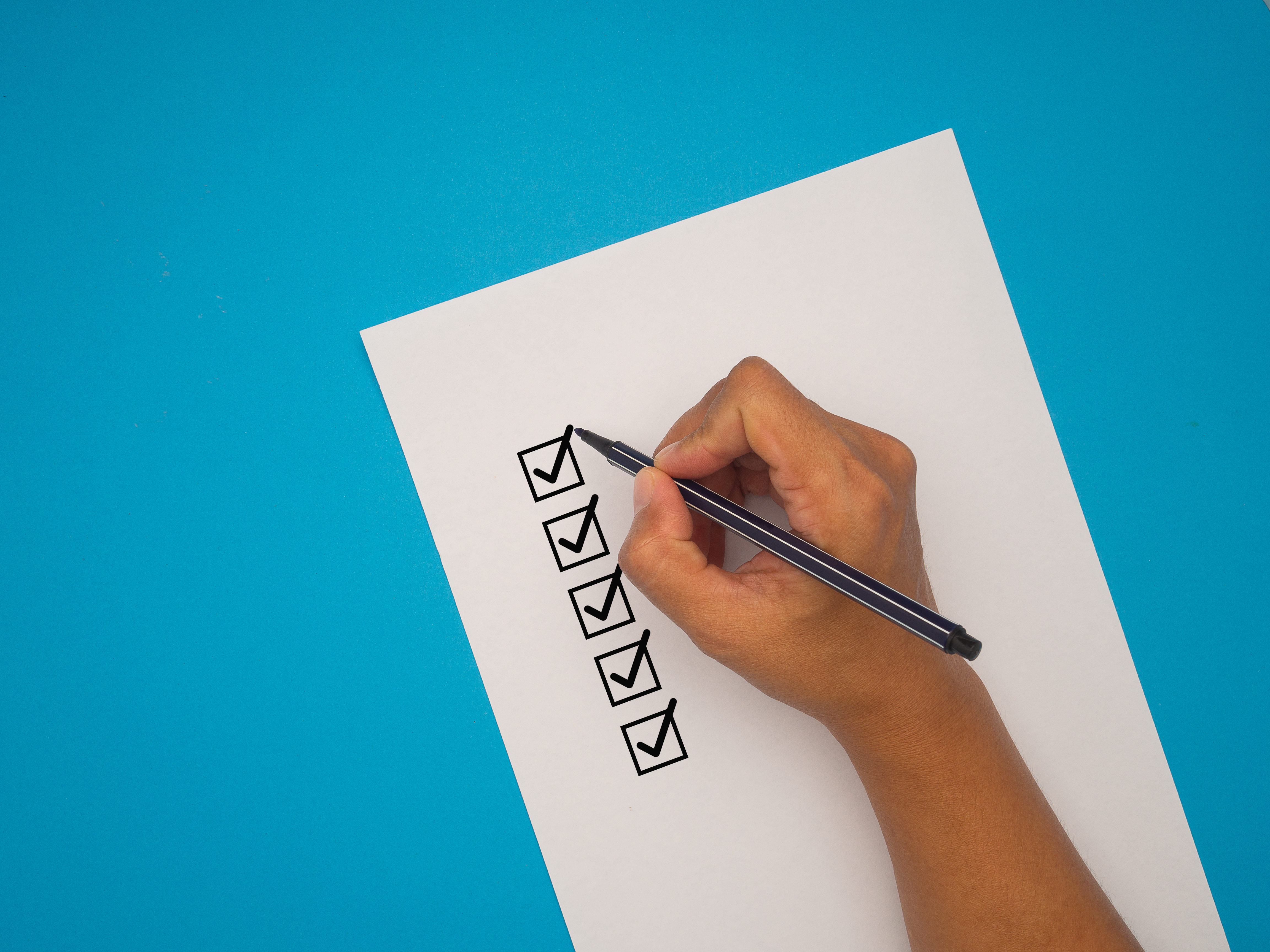 Checklist concept. Close-up of a hand holding a pen, mark five correct sign symbols in boxes on white paper over a blue background. Space for text. Checklist, Approved concept