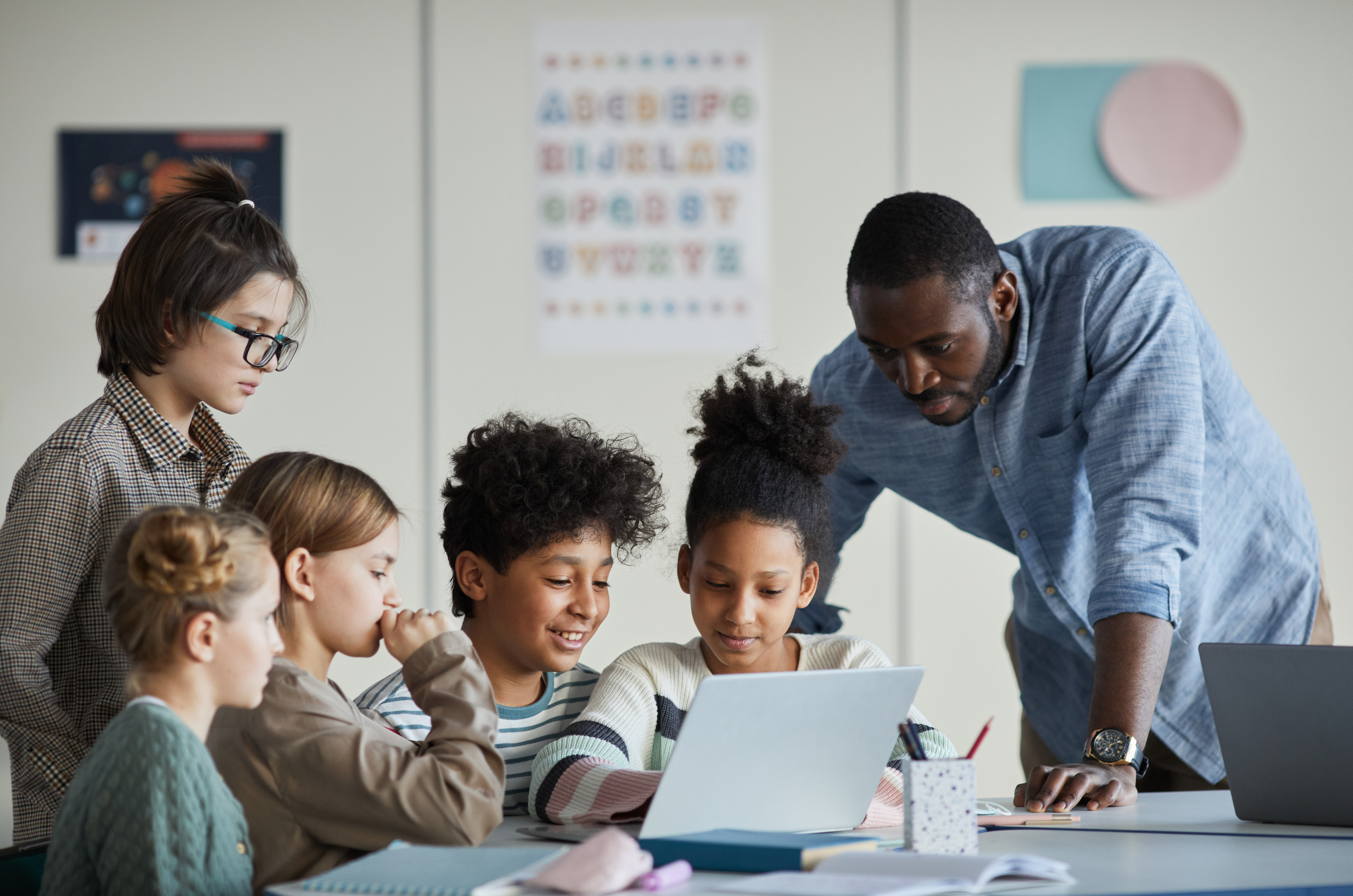 Diverse group of children with male teacher using laptop together in modern school classroom