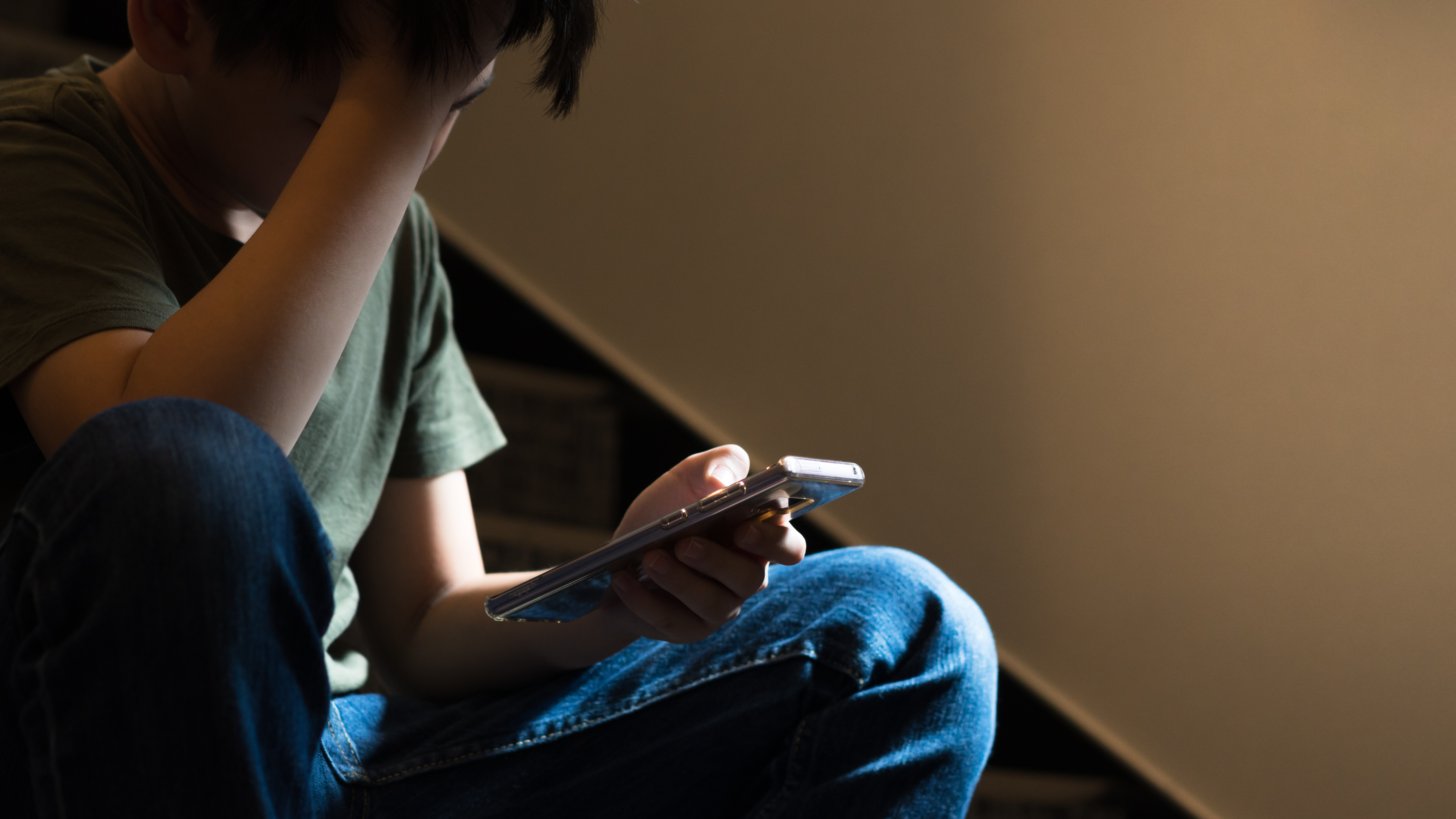 Young Asian preteen/teenage boy sit on stair at home, cry and cover his face with hand, other hand hold smartphone, feeling alone, stressed, frustrated, overwhelmed, depressed. Cyber bullying concept.
