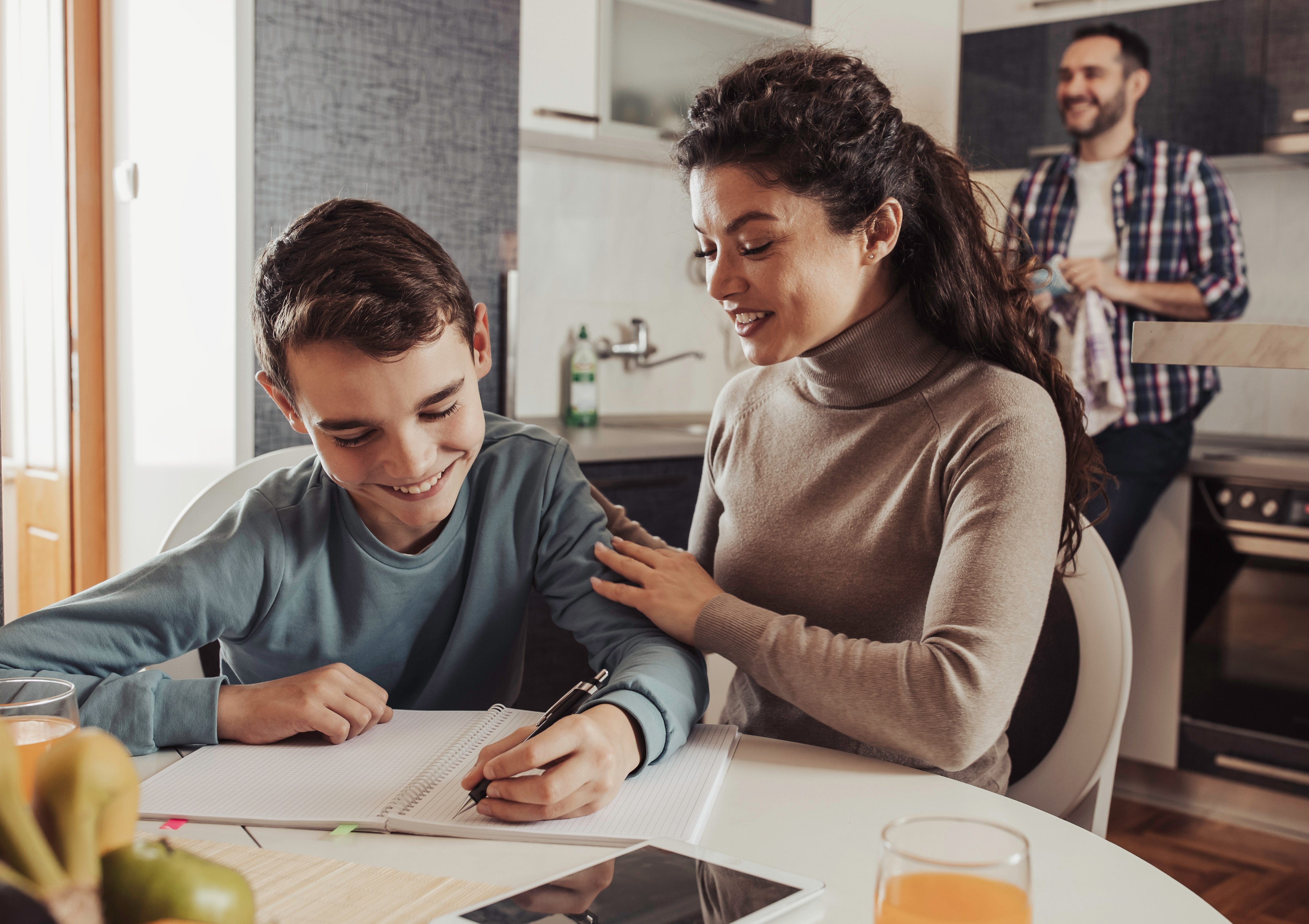 Mother sitting with son while doing homework on table in kitchen, smiling, happy, satisfied expression. Father doing chores in background