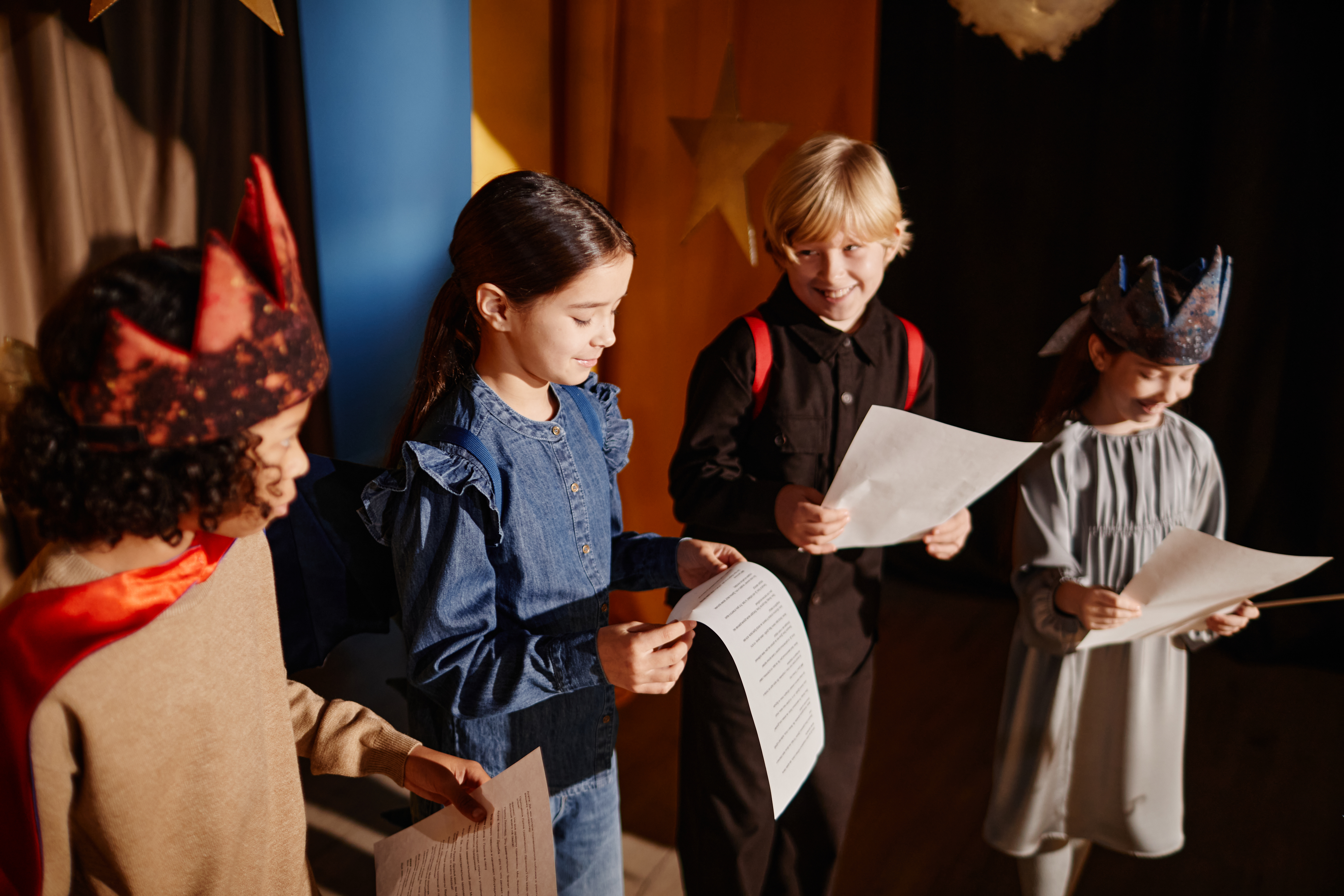 Young students rehearsing for school play, some wearing costumes and holding scripts, happily engaging in theater activity. Wooden floor and colorful backdrop create vibrant atmosphere