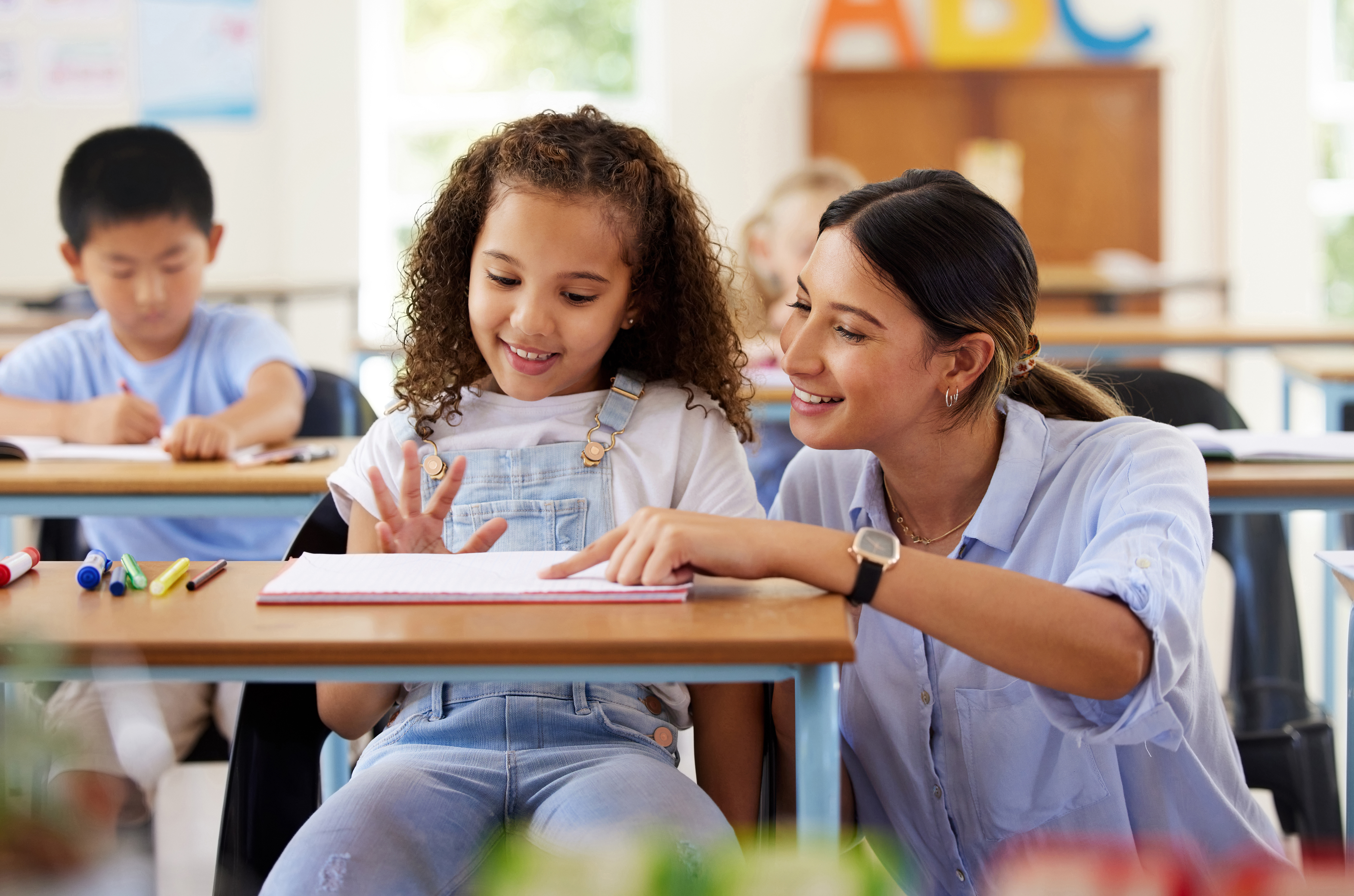 Teacher, learning and helping girl in classroom for knowledge, studying and assessment. Preschool smile, development and happy kid or student with woman for education with notebook in kindergarten