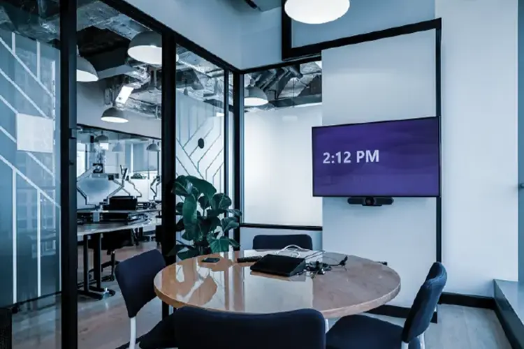 Glass-walled Microsoft Teams meeting room with a round conference table and wall-mounted display.